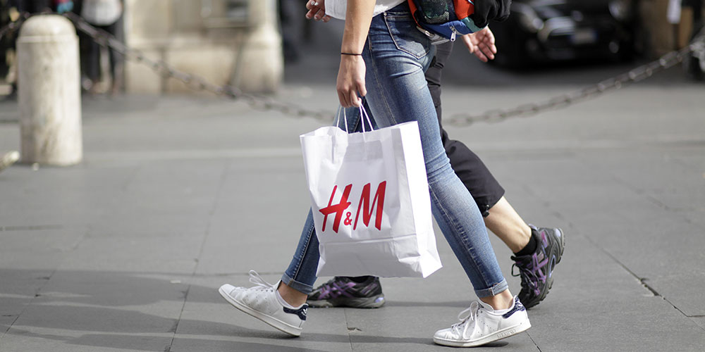 The legs of two women walking down a sidewalk holding a full H&M bag.