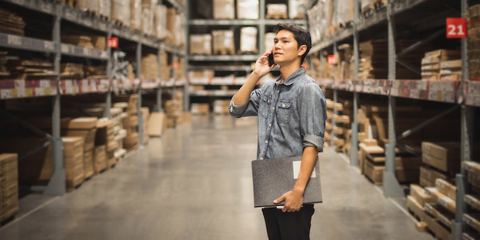 Person standing in the aisle of a warehouse, holding a clipboard while talking on the phone.