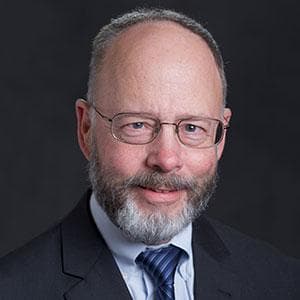 Headshot of Jim Reeve - bearded man, wearing wire-rimmed glasses and a dark suit and blue tie.
