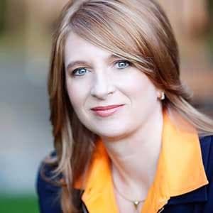 Headshot of Kate Vitasek - woman looking at the camera, wearing an orange shirt and a dark suit jacket.