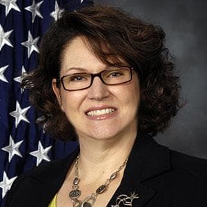 Headshot of Pam Donovan - woman with shoulder length curly hair, dark-rimmed glasses and a dark suit jacket standing in front of an American flag.