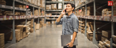 Person standing in the aisle of a warehouse, holding a clipboard while talking on the phone.