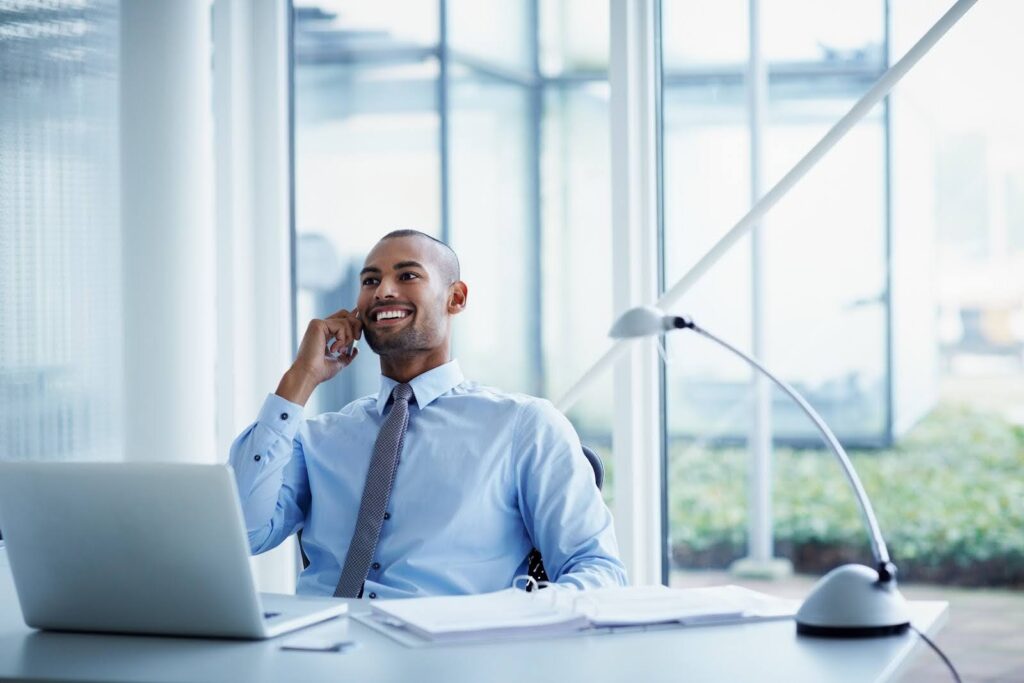 A smiling business person seated at a desk in a private office uses a mobile phone.