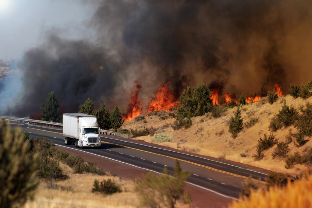 A semi-truck drives down a highway beside a wildfire.