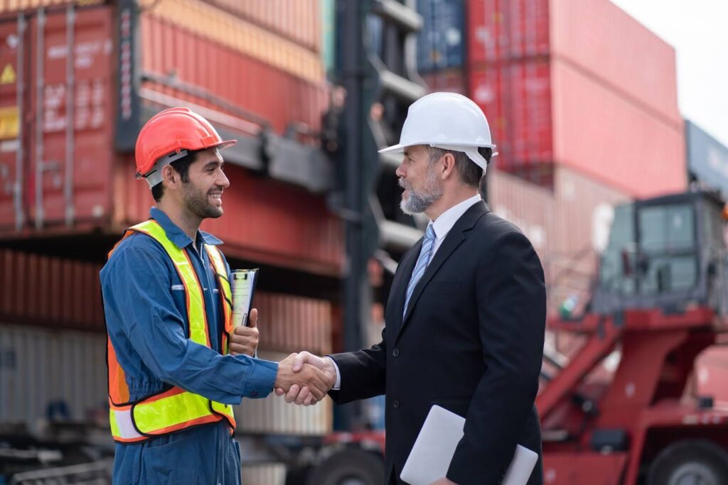 A supply chain executive and a dock supervisor standing in front of a container ship shake hands.