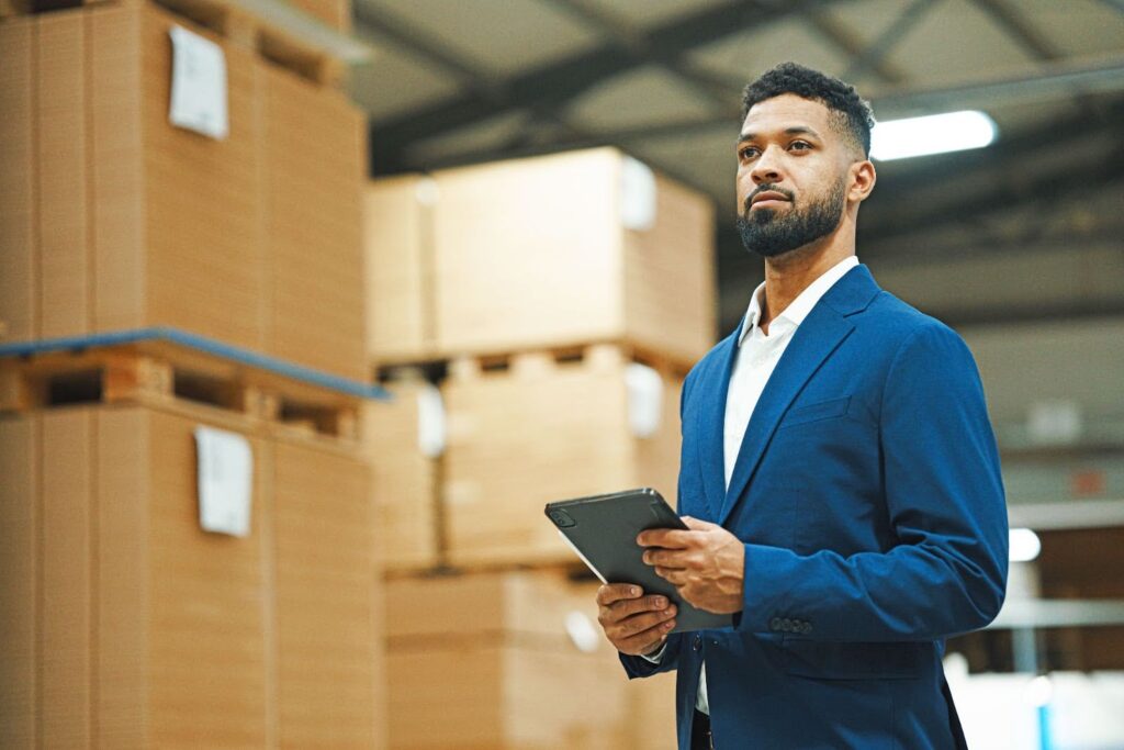 A logistics professional in business attire surveys a warehouse; he is holding a tablet.